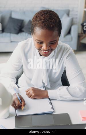 Smiling black businesswoman working on laptop and taking notes in modern office. Young afro-american woman. Freelancer using laptop, phone and the Stock Photo
