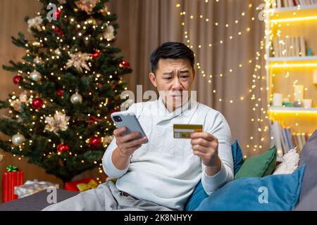 Dissatisfied Asian man trying to make a purchase in an online internet store, man cheated on Christmas, sitting on sofa in living room with smartphone in hands and credit card at home near Christmas tree. Stock Photo