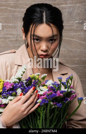 Portrait of brunette asian woman in trench coat holding flowers on abstract brown background Stock Photo