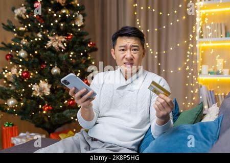 Cheated man at home sitting on sofa and looking at camera, Asian Christmas day frustrated trying to make a purchase in online store and bank transaction celebrating new year . Stock Photo