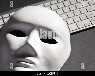 White face mask and computer keyboard on black background. Hacker, computer hacking, cybercrime, scam or online security concept. Stock Photo