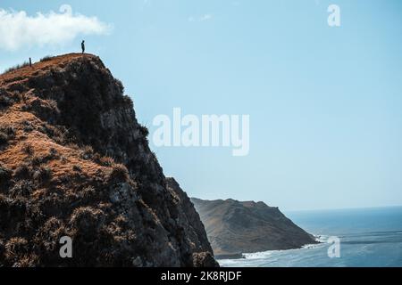 A man standing at the edge of a tall cliff near a sea with a blue sky in the background Stock Photo