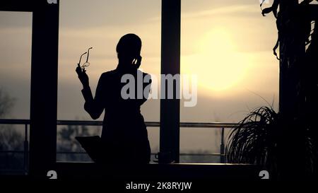 dark silhouette of Young business woman. she holds glasses in her hand, talks emotionally on mobile phone, gesticulates with hands, against background of large office window, at sunset, in rays of light. High quality photo Stock Photo