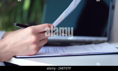 Cropped shot of woman's hand writing something while using laptop at desk. close up, on a white desktop there is a white cup of tea, next to a laptop and spread out business graphs. High quality photo Stock Photo