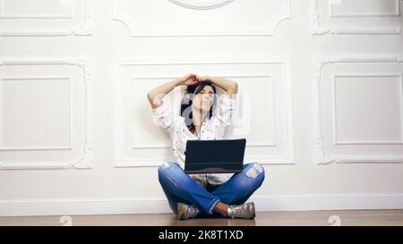 young woman, girl, brunette, in white shirt and jeans, listening to music, using headphones and laptop while sitting with legs crossed on the floor over white wall with decorative stucco,. High quality photo Stock Photo