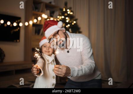 Focus on sparklers, blurred happy father and little cute daughter holding bengal fire in their hands on background of decorated christmas tree at home. Stock Photo