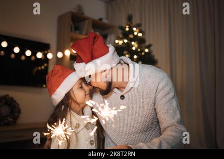 Family, winter holidays and people concept. Portrait of happy father and little cute daughter holding sparklers in their hands on background of decorated christmas tree at home. Stock Photo