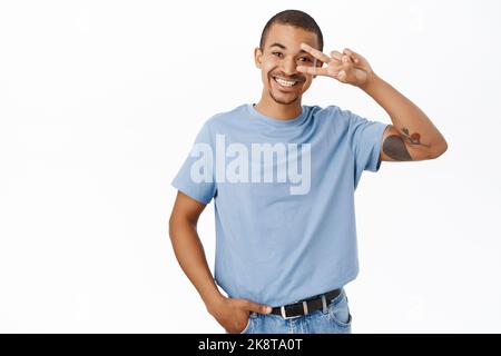 Positive guy shows peace, v-sign gesture and smiles, stands in casual clothes against studio background Stock Photo