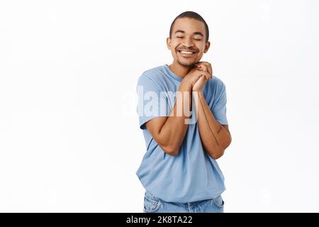 Dreamy young man hold hands together, eyes closed, smiling while dreaming, thinking of smth cute, standing over white background Stock Photo