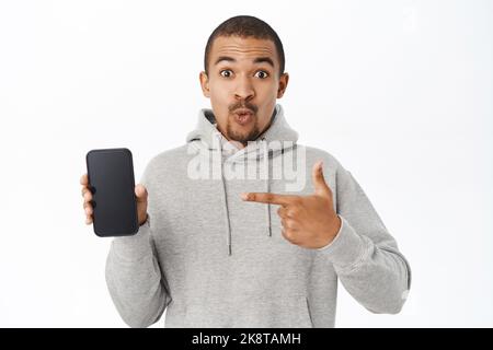 Handsome man shows mobile phone screen, points at telephone and smiles, recommends application, stands over white background Stock Photo