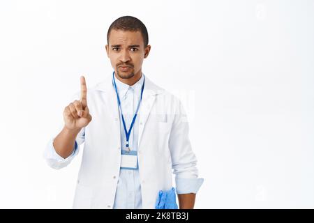 Serious doctor shows stop gesture, one finger, talking to patient, standing concerned over white background Stock Photo