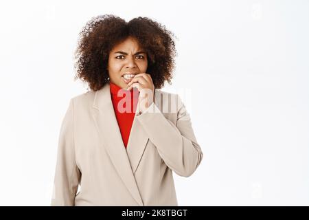 Anxious female office worker, businesswoman bites finger and looks worried, feeling nervous, standing over white background Stock Photo