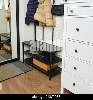 An entrance hall with a hanger and a chest of drawers in a home apartment. Shoes on shelves and jackets on hangers in the hallway by the front door wi Stock Photo