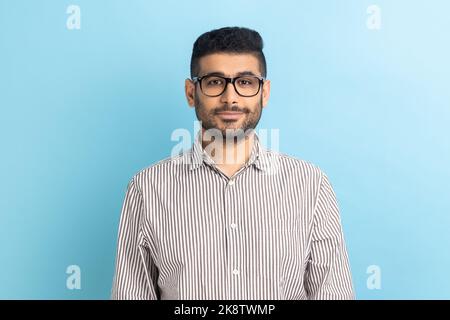 Portrait of smiling positive pleased businessman in optical eyeglasses standing looking at camera with smile, wearing striped shirt. Indoor studio shot isolated on blue background. Stock Photo
