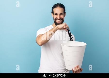 Portrait of man with beard wearing white T-shirt throwing out eyeglasses into rubbish bin, treat his eyesight, looking smiling at camera. Indoor studio shot isolated on blue background. Stock Photo