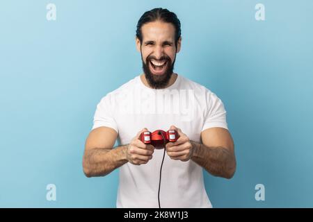 Portrait of man with beard wearing white T-shirt holding in hands red gamepad joystick, looking at camera with optimistic expression, enjoying game. Indoor studio shot isolated on blue background. Stock Photo