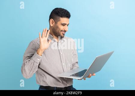 Businessman waving hand gesturing hello to laptop screen, talking on video call with coworker, communicating online conference, wearing striped shirt. Indoor studio shot isolated on blue background. Stock Photo