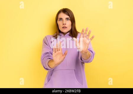 No, it's scary. Portrait of frightened shocked woman raising hands in fear, looking horrified and panicking, hiding from phobia, wearing purple hoodie. Indoor studio shot isolated on yellow background Stock Photo