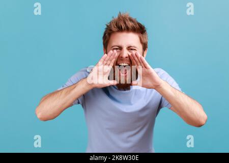 Portrait of angry nervous bearded man loudly yelling widely opening mouth holding hands on face, screaming announcing his opinion. Indoor studio shot isolated on blue background. Stock Photo