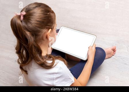 Little girl sitting on the floor and holding a white tablet with isolated screen for promoting video games, websites or apps Stock Photo
