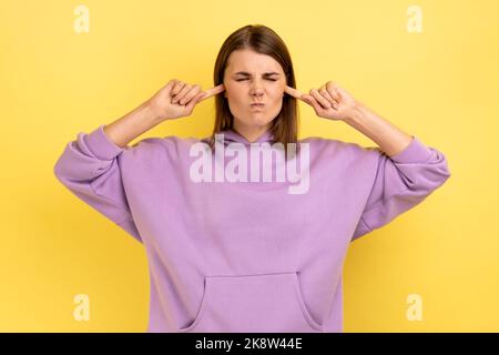 Don't want to listen. Beautiful irritated stressed out woman clothing covering ears, keeping eyes closed, nervous breakdown, wearing purple hoodie. Indoor studio shot isolated on yellow background. Stock Photo