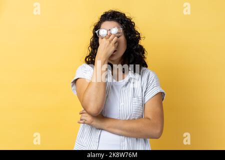 Portrait of tired exhausted attractive young adult woman with dark wavy hair standing with raised eyeglasses, feels pain in eyes. Indoor studio shot isolated on yellow background. Stock Photo