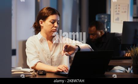 Young adult checking time on wristwatch to leave office after hours, working at night on research data to create presentation. Feeling overworked at corporate job, planning financial report. Stock Photo