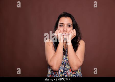 Sad indian woman holding clenched fists on cheeks portrait, cute pose. Calm lady touching face with hands, young person standing with neutral facial expression, looking at camera Stock Photo