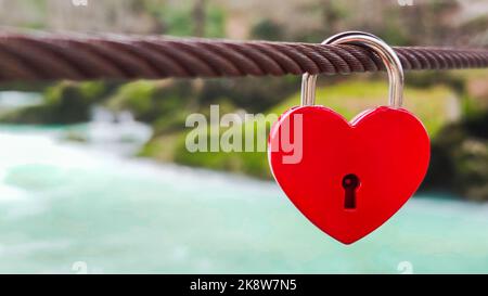 Castle in shape of red heart hangs on metal cable against background of river and nature. Concept of love and health. Close-up, copy space Stock Photo