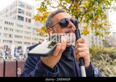 Blind middle-aged man with gray hair and beard wearing sunglasses holding cane and phone using text-to-speech function. Accessibilty. Modern technology. Horizontal outdoor shot. High quality photo Stock Photo