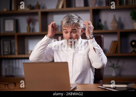 Happy businessman surprised by the good news and making yes gesture while looking at laptop sitting at office. Trader enjoying success, celebrating Stock Photo