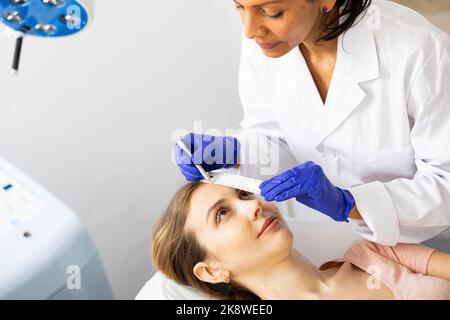 Beautician using white pencil and ruler for marking skin of young woman Stock Photo