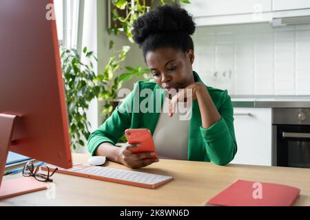 Focused African American woman holding phone reading business press sits at computer desk in office Stock Photo