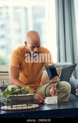 My doctor said that I should monitor my blood pressure regularly. a young man taking his blood pressure while sitting on the sofa at home. Stock Photo