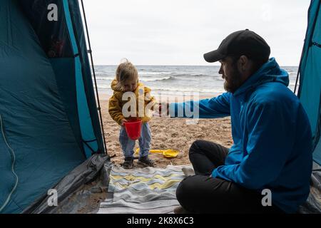 Dad and baby are playing together in a tent on the seashore. The concept of adventure, family camping, outdoor activities, bonding. Stock Photo