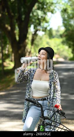 Middle age woman resting and drinking water from bottle during cycling in summer park. Retirement people lifestyle concept Stock Photo
