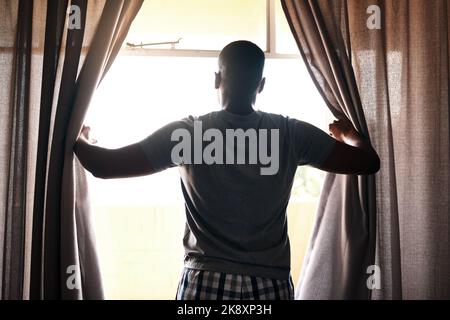 Im going to seize the day. Rearview shot of an unrecognizable man standing alone in his bedroom and opening his curtains in the morning. Stock Photo