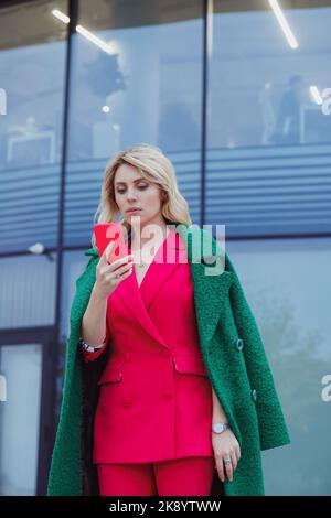 mature blonde curly woman in suit walks in business district and uses her phone. Pretty autumn woman in pink suit walks down the street looking at her Stock Photo