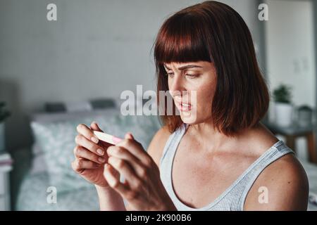 Life is full of unexpected surprises. an attractive young woman looking shocked while looking at her pregnancy test results at home. Stock Photo