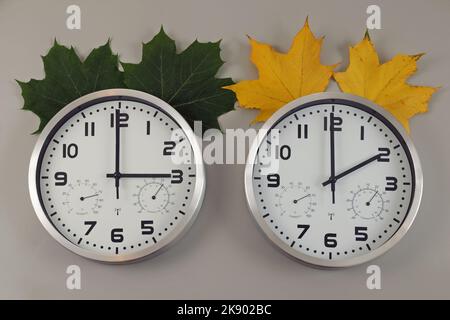 Two clocks, one shows two o'clock, the other shows three o'clock. Symbol of time change. Green leaves for summertime, yellow for wintertime. Stock Photo