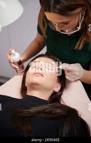 Close up of beauty specialist in sterile gloves applying cleansing mousse on woman eyelashes with cosmetic lash brush. Eyelash stylist preparing woman lashes for applying extensions. Stock Photo
