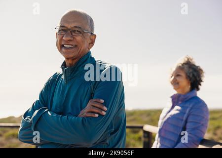 Happy senior man smiling at the camera while standing on a foot bridge with his arms crossed. Cheerful elderly man enjoying a relaxing seaside holiday Stock Photo