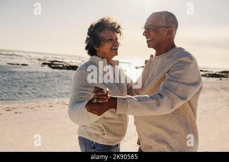 Senior couple smiling while dancing together at the beach. Happy elderly couple having a good time next to the ocean. Cheerful mature couple enjoying Stock Photo