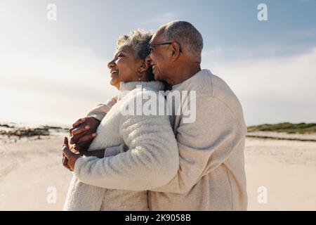 Senior couple enjoying a refreshing view of the ocean at the beach. Happy elderly couple embracing each other while standing on beach sand. Mature cou Stock Photo