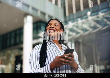 Thoughtful young businesswoman looking away with a smile while commuting to work in the city. Happy young business woman using a smartphone on her way Stock Photo