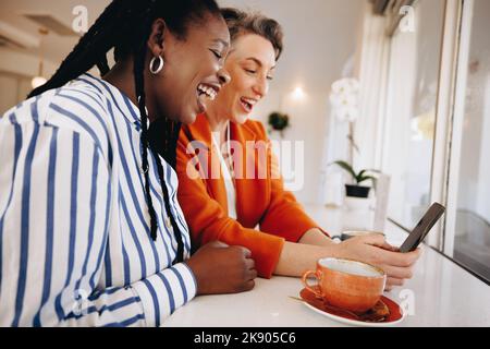 Two happy business women video calling their team on a smartphone in a coffee shop. Smiling businesswomen having a virtual morning catch up with their Stock Photo