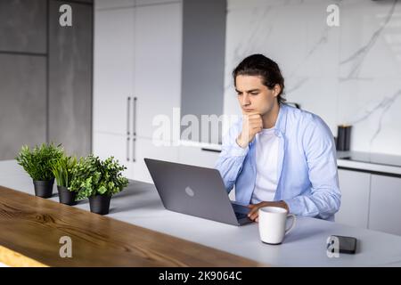 Serious focused determined young bearded businessman or freelancer in glasses and casual clothes sitting at kitchen table, typing on laptop, working o Stock Photo