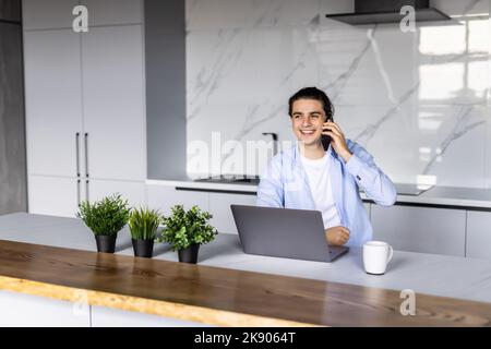 Young positive man freelancer working remotely at home, looking at laptop and consulting client via cellphone, sitting at kitchen Stock Photo