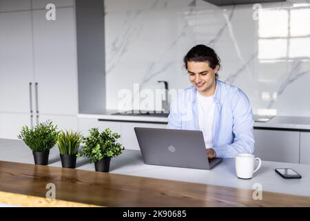 Smiling young man working on laptop in modern kitchen, checking email in morning, writing message in social network, happy young male using internet b Stock Photo