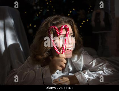 beautiful adult girl of 18 years old lies near Christmas tree , holds two red lollipops in front of face, folded in shape of heart, smiles cheerfully, Stock Photo